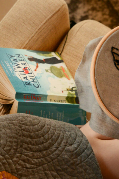 image of a child sitting on a sofa with a copy of The Railway Children cracked open with the pages down and the spine and cover facing up. The child has switched from reading to working on an embroidery hoop with in the pattern of a bee.