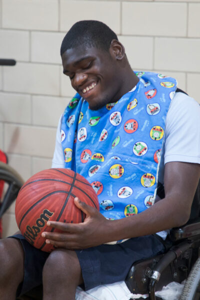 A teen boy in a wheelchair holding and exploring a basketball inside a gym.