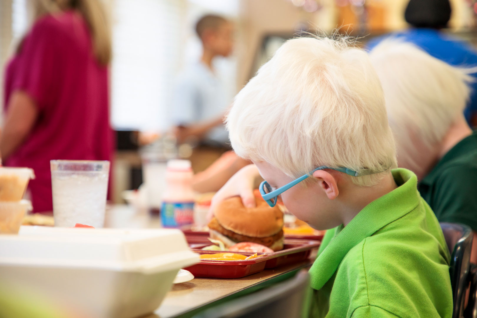 A young boy wearing glasses sitting at a cafeteria table eating.