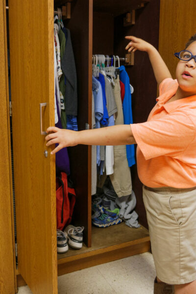 A young boy opening a closet pointing to his clothing. 