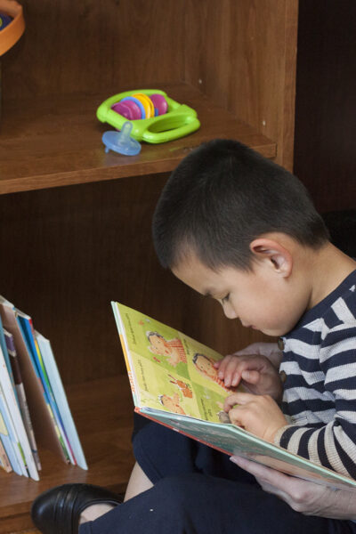 A toddler boy reading a book and feeling braille while sitting on his mom's lap near a book case.