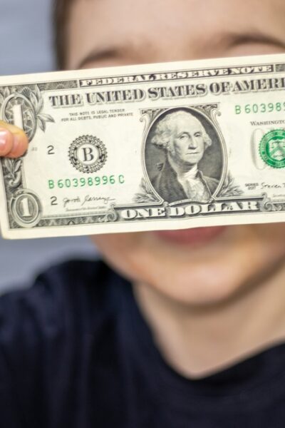 One dollar bill in front of kid child boy face cover eyes or mouth.