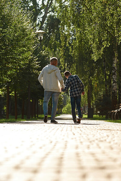 Back view of caucasian father and teenage son with cerebral palsy walking on pavement in sunny park.