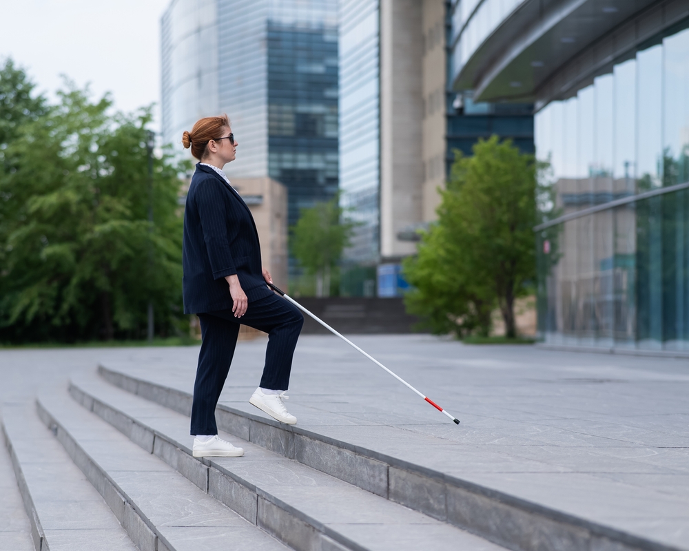 Person wearing business attire and holding a white cane walks up the steps to an office building.